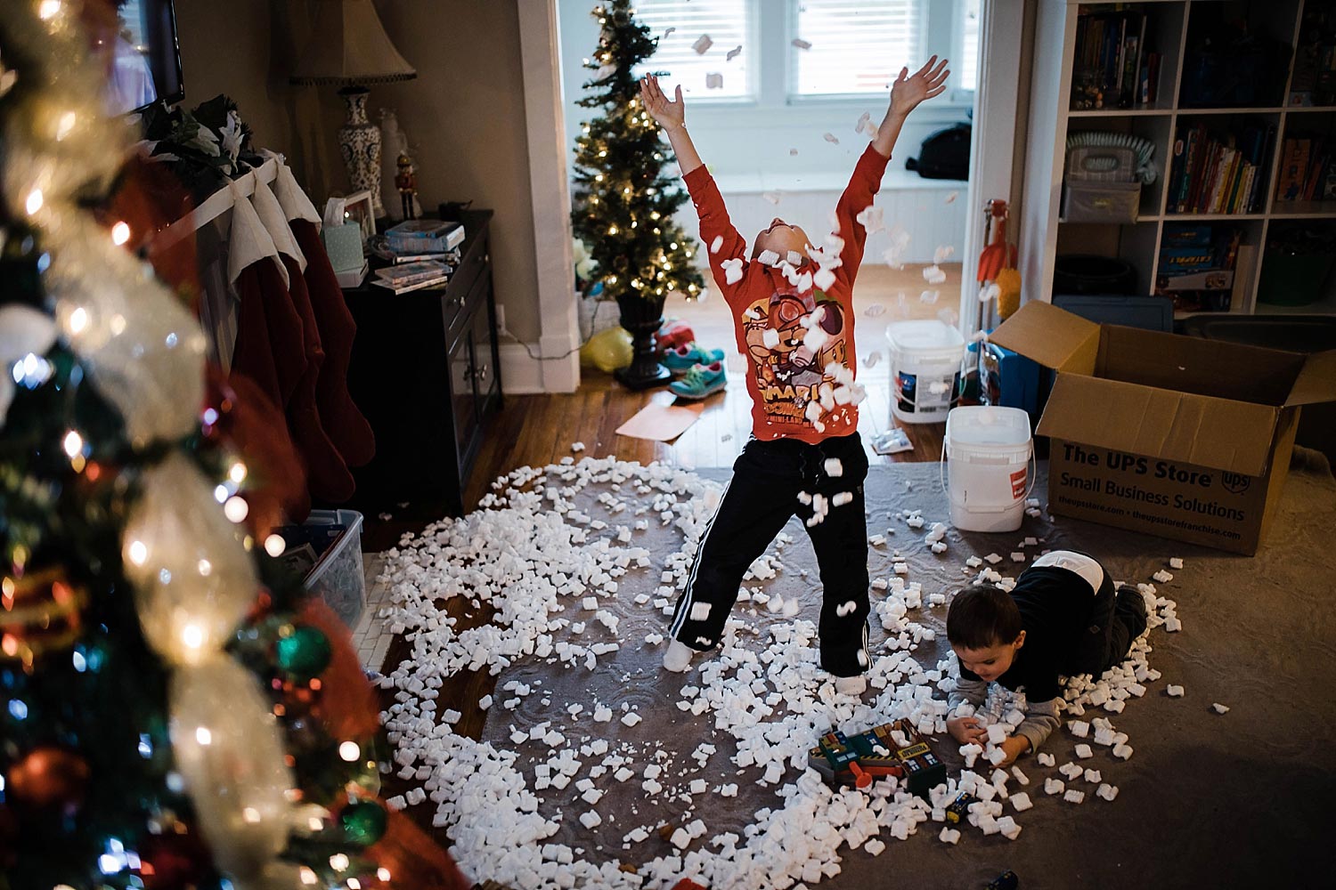boy throws packaging peanuts into air next to Christmas tree