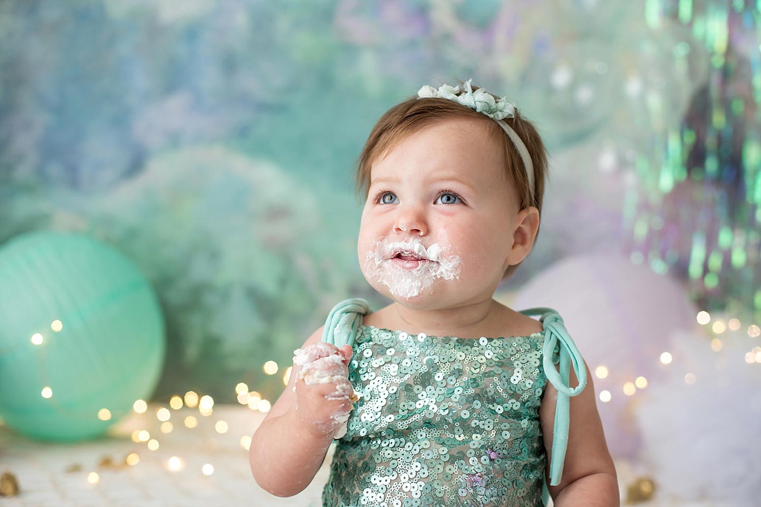 One year old baby girl in mermaid themed set smirks with cake icing all over her face