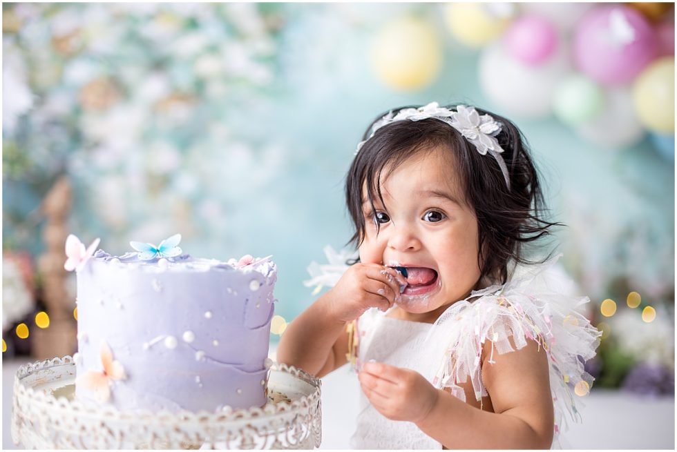 baby in white dress bites into pastel butterfly cake